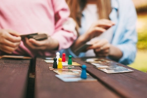 Twee mensen spelen buiten een bordspel, waarbij de aandacht vooral uitgaat naar de kleurrijke speelstukken. De sfeer is ontspannen en boeiend, en straalt zowel ontspanning als strategisch spel uit.